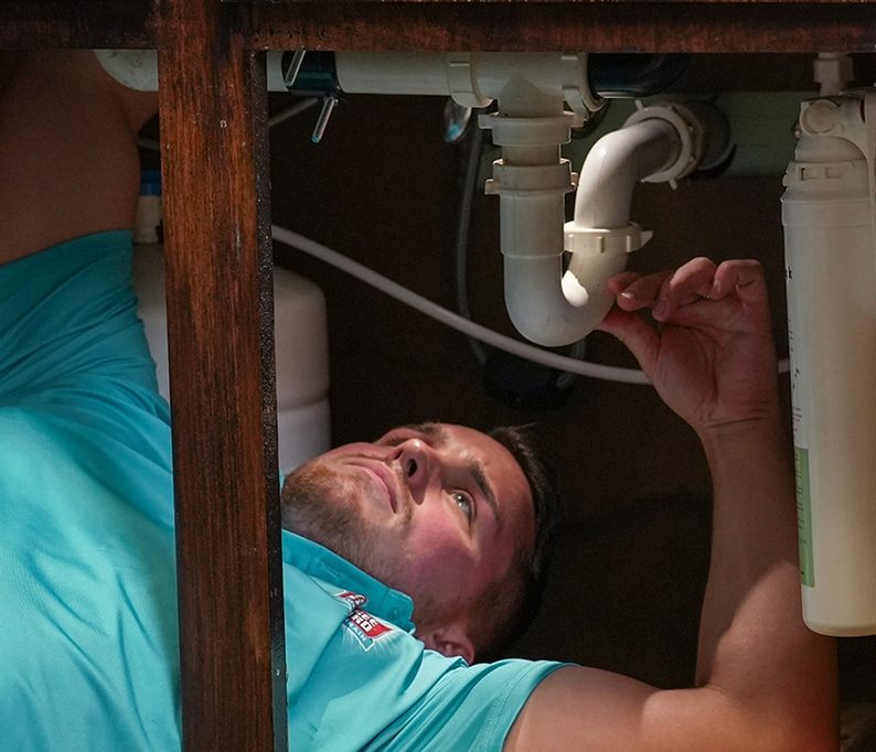 A man wearing a teal shirt lies under a sink, inspecting and working on the white plumbing pipes.