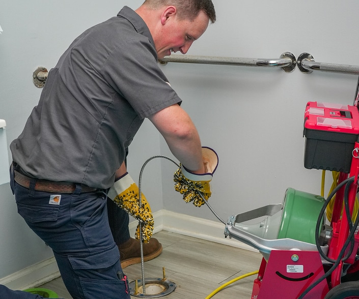 A plumber uses a drain snake machine to clear a clog from a floor drain in a restroom.