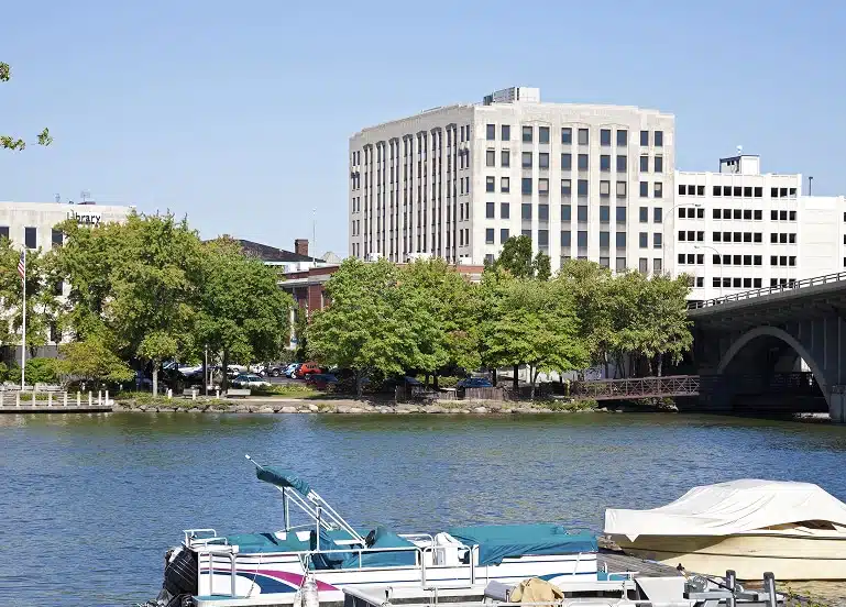Boats docked along a river with trees and several mid-rise office buildings in the background under a clear blue sky.
