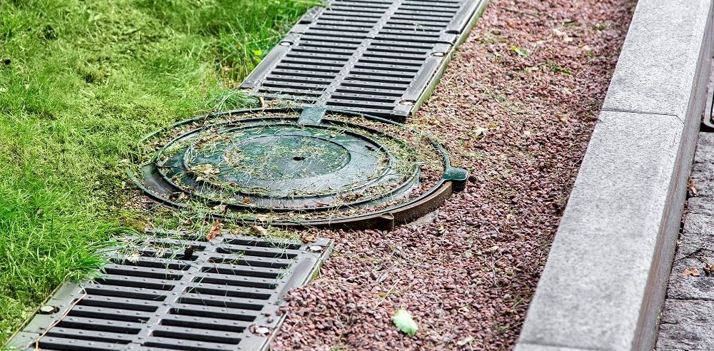 A metal manhole cover and drainage grates, part of Drainage Systems in Knoxville, are set in the ground next to a curb, with grass and gravel surrounding them.