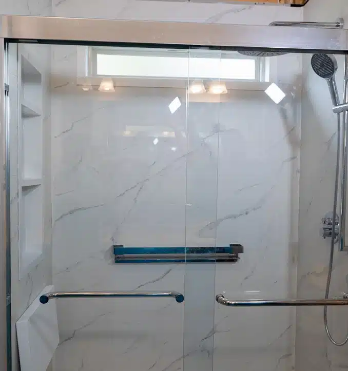 Modern walk-in shower with white marble-style tile walls, glass sliding doors, chrome grab bars, a handheld showerhead, and built-in shelving niche.