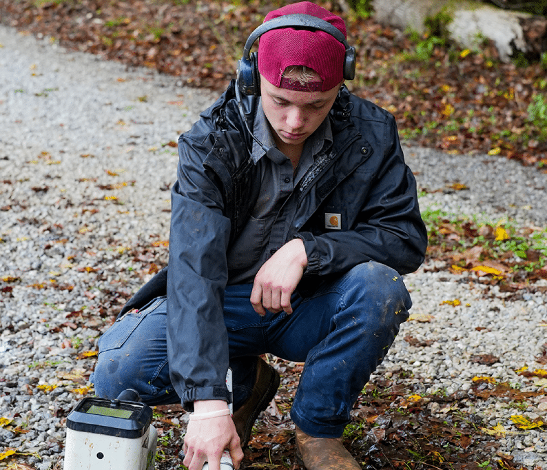 A person wearing a red cap and headphones squats on a gravel path, focusing intently on a device in front of him