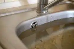 A stainless steel kitchen sink filled with dirty, cloudy water and food debris, nearly reaching the overflow drain.