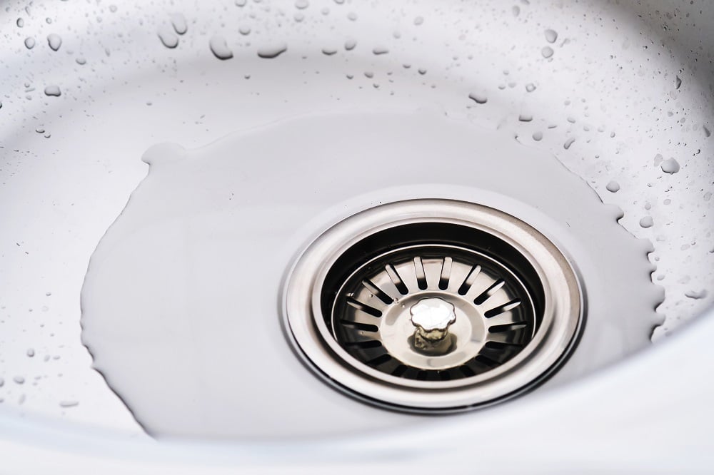 A close-up of a stainless steel sink drain with water pooling around it and droplets visible on the sink’s surface, highlighting the need for commercial drain cleaning.