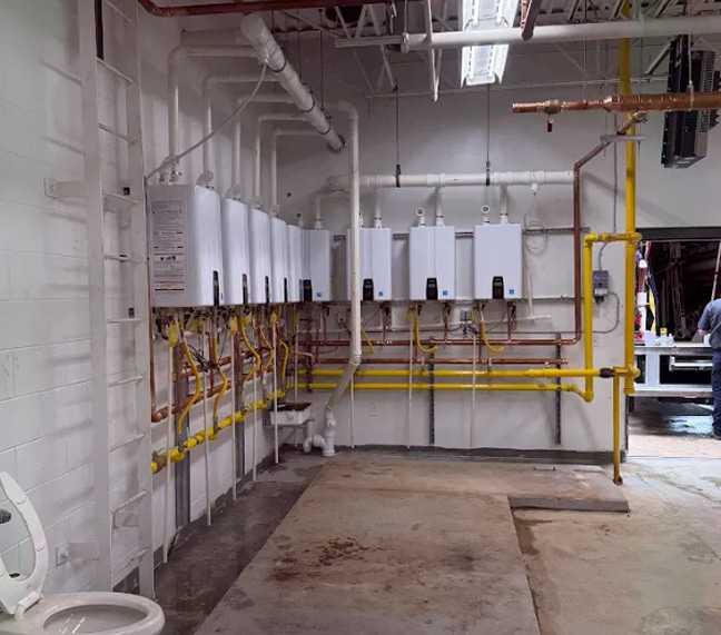 A row of wall-mounted tankless water heaters with connected pipes and valves in a utility room with exposed ceiling fixtures.