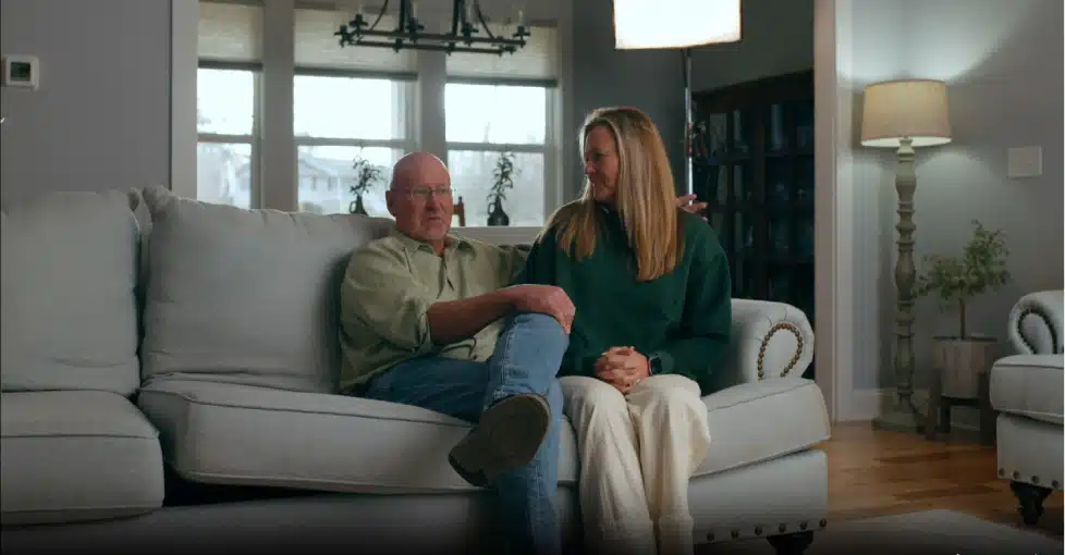 Two people sit together on a light-colored couch in a living room, with large windows and a lamp in the background.