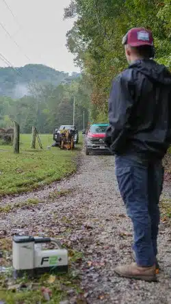 A person stands on a gravel road watching a utility crew perform water line service with machinery near a red truck and open field lined with trees and power lines.