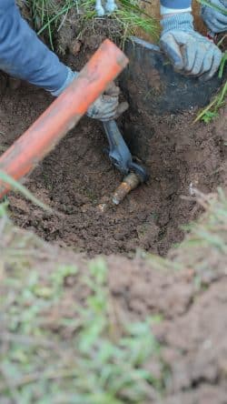 A person wearing gloves uses a tool to dig around and expose a metal pipe in a dirt hole.