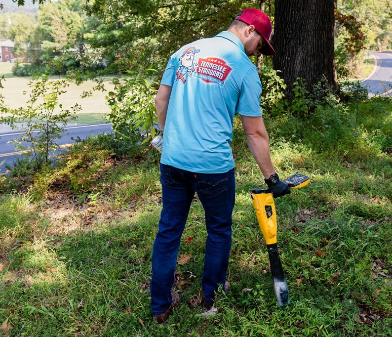A man wearing a blue T-shirt and red cap uses a yellow metal detector on a grassy area near trees.