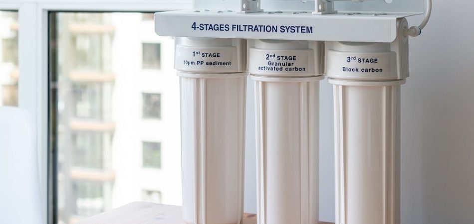 Four stages water filtration system for the home with: coconut postcarbon filter, granular activated and block carbon on a wooden table against a blurred window background. Reverse osmosis system