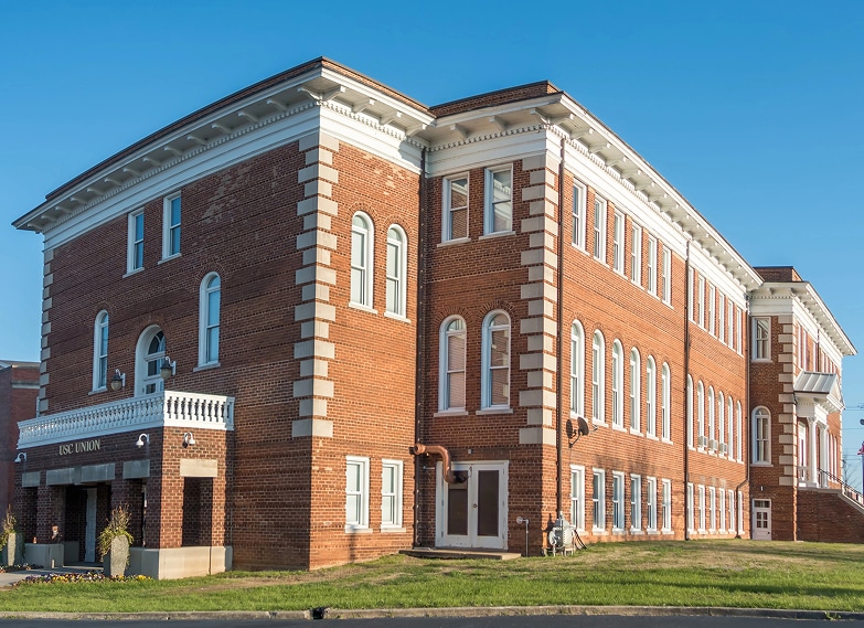 A three-story red brick building with white trim and arched windows, featuring a small entrance portico on the left side, under a clear blue sky.