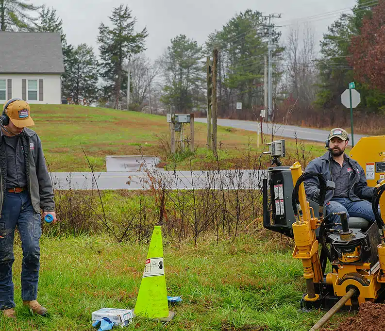 Two workers in jackets and hats operate machinery near a roadside, with a yellow caution cone and equipment on the grass in a rural area.