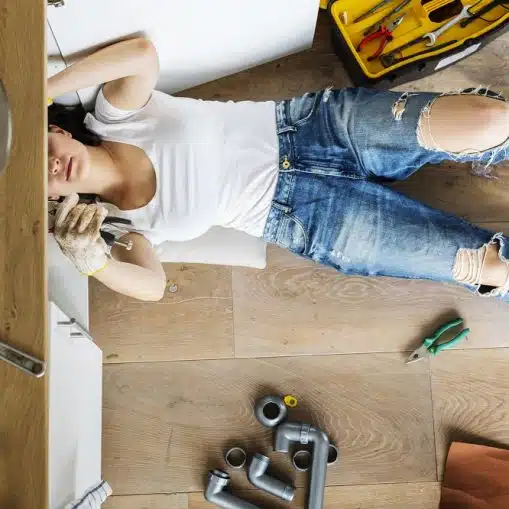 A person in ripped jeans and a white shirt lies on the floor fixing pipes under a kitchen sink, surrounded by plumbing tools and parts.