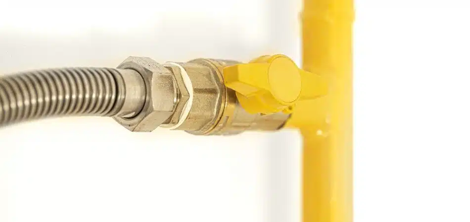 Close-up of a yellow gas valve connected to a metal pipe and flexible hose, part of a gas line for an outdoor kitchen in Maryville, mounted on a white wall.